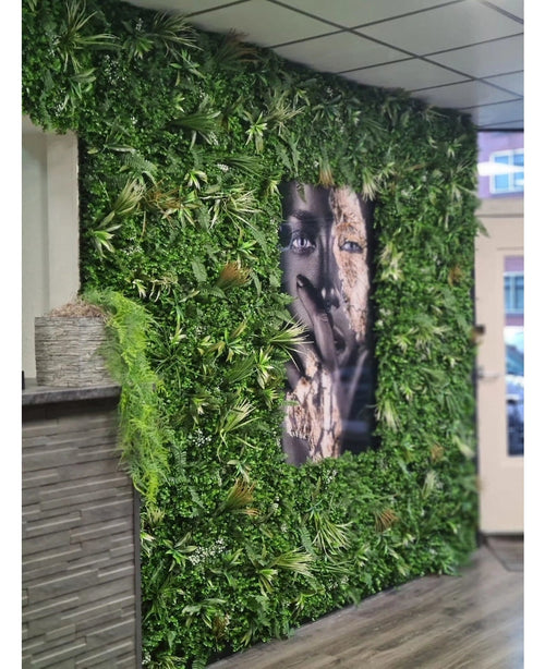 Una pared cubierta con un seto artificial Savanne de 100x100 cm, con vegetación protegida contra los rayos UV en una maceta de plástico de Ø100, tiene en el centro un gran retrato en blanco y negro. El interior moderno tiene un techo de tejas y un mostrador de madera a la izquierda.