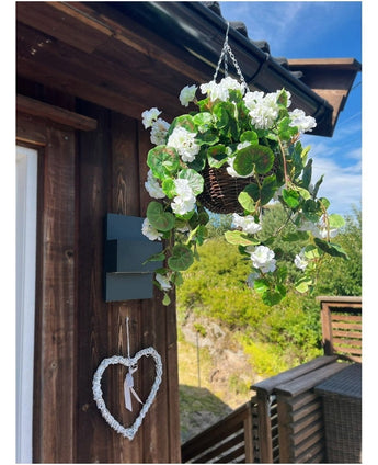 De Kunst begonia Wit in Mand UV 70cm hangt aan een houten muur, met daaronder een decoratief wit hart. Groen, een houten vlonder en een gedeeltelijk bewolkte blauwe lucht maken de achtergrond compleet.