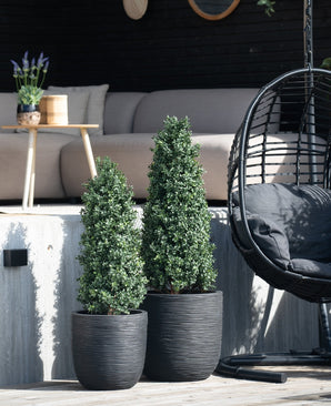 Dos plantas artificiales de boj piramidal UV de 120 cm se colocan sobre una terraza de madera frente a una zona de estar al aire libre con un sofá luminoso, una mesa redonda con lavanda y una silla colgante negra.