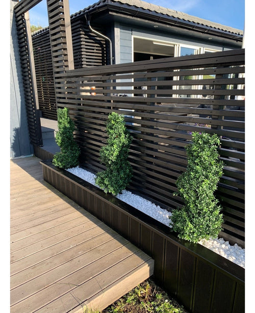 Tres arbustos topiarios artificiales en espiral de boj estilo romano UV de 90 cm se encuentran en un estrecho lecho de guijarros blancos, contra una moderna valla de privacidad de listones negros junto a un patio de madera y una casa contemporánea.