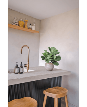 Una encimera moderna con grifo de latón, dos dispensadores de jabón, la planta en maceta Planta de Tabaco Artificial Hoja de Violín UV de 45 cm y dos taburetes de madera. En una pared de color claro hay un estante flotante con tarros y botellas.