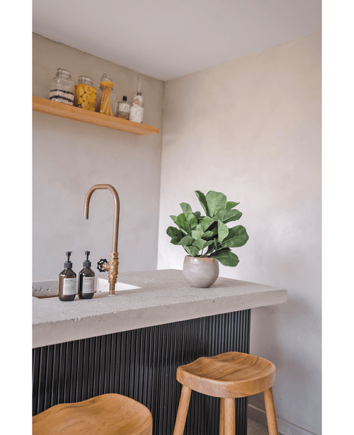 Una encimera moderna con grifo de latón, dos dispensadores de jabón, la planta en maceta Planta de Tabaco Artificial Hoja de Violín UV de 45 cm y dos taburetes de madera. En una pared de color claro hay un estante flotante con tarros y botellas.