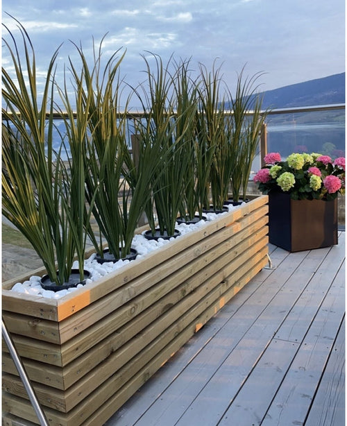 Una jardinera de madera con Césped Artificial Verde UV 90cm y piedras blancas se alza sobre un deck de madera junto a una jardinera negra con hortensias de colores, con balaustradas de cristal y una vista de las montañas al fondo.