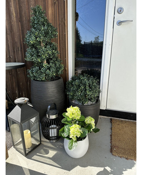 En la terraza, cerca de una puerta marrón, hay una planta artificial de hortensia verde UV de 35 cm en una maceta blanca con flores de color amarillo verdoso, junto a dos plantas en una maceta negra y dos faroles decorativos con velas.