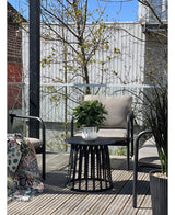 Een modern terras met twee stoelen met kussens, een ronde zwarte tafel met de Kunstplant Laurier UV d33 cm als middelpunt en een deken met patroon op één stoel. Bomen en een houten hek verschijnen op de achtergrond onder een heldere hemel.