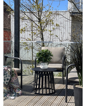 Una terraza moderna con dos sillas con cojines, una mesa redonda negra con la planta Laurel Artificial UV d33 cm como centro de mesa y una manta estampada sobre una silla. Al fondo aparecen árboles y una valla de madera bajo un cielo despejado.