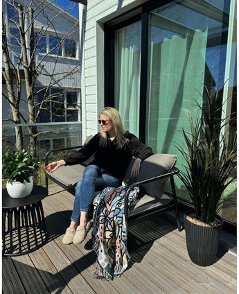 Een vrouw met zonnebril, zwart topje en blauwe jeans ontspant op een kussensstoel op een zonnig balkon met een drankje in de hand. De ruimte is voorzien van de Kunstplant Laurier UV d33 cm, een levendig decor, een kleurrijke deken en een kleine tafel met planten.