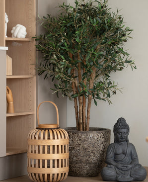 Un olivo artificial UV de 120 cm se encuentra junto a una estatua de Buda gris y una linterna de madera en el suelo, junto a una tabla de madera clara con adornos en forma de manos. Esta planta de bajo mantenimiento aporta una atmósfera tranquila y serena a la habitación.