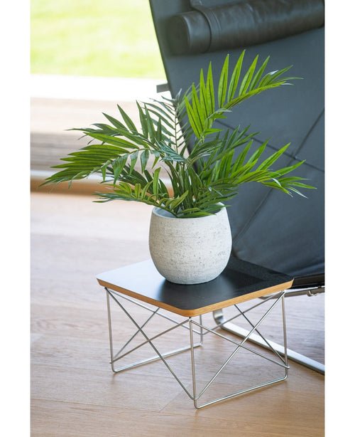 Una palmera artificial UV de 60 cm se encuentra en una maceta sobre una pequeña mesa auxiliar moderna con tablero negro y patas de alambre metálico, junto a una silla negra sobre un suelo de madera clara.