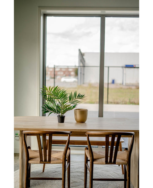 Una mesa de madera con dos sillas, una palmera artificial UV de 60 cm en una maceta y un jarrón dorado se colocan cerca de un gran ventanal que da a una zona industrial con edificios y una valla.