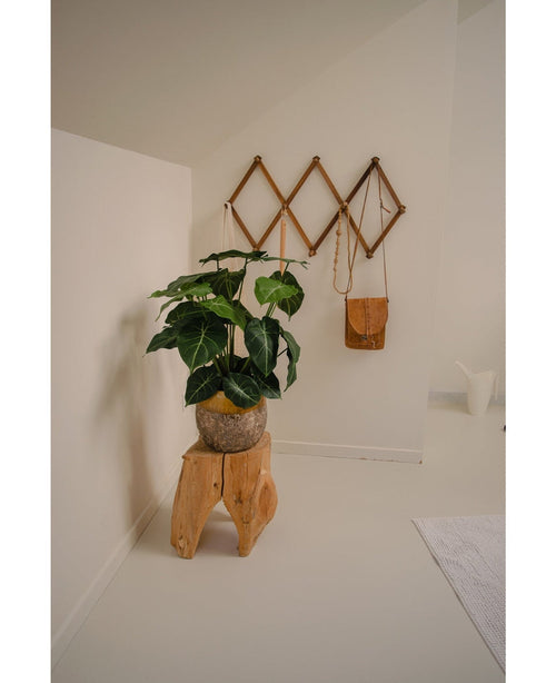 Un rincón de una habitación minimalista se convierte en un oasis verde con la Planta Artificial Syngonium UV de 58 cm sobre un taburete de madera natural, junto a un bolso de cuero marrón sobre un estante de pared geométrico y una jarra blanca, todo ello contra las paredes y el suelo de color blanquecino.