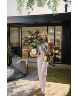 Een glimlachende vrouw staat op een houten dek buiten een modern huis, met twee Kunstplant Calathea Makoyana UV 60 cm rood met kleurrijke bladeren. Ze draagt een lichtroze broek en een wit topje; buitenmeubilair is zichtbaar op de achtergrond.