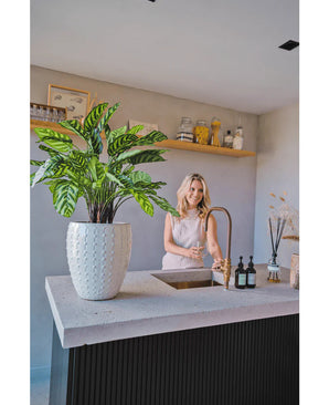 Una mujer sonríe ante la encimera de una cocina moderna con la mano bajo el grifo. La Planta Artificial Calathea Makoyana Deluxe UV 75 cm está en primer plano sobre el mostrador. Los estantes abiertos exhiben macetas y artículos decorativos al fondo.