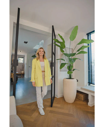 Een glimlachende vrouw in een gele blazer staat in een lichte, moderne kamer met houten vloeren, naast een Kunstplant Strelitzia 210 cm real touch bij de glazen deur. Zonlicht filtert door de jaloezieën aan de rechterkant.