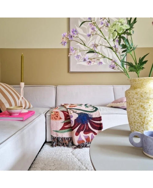 Una acogedora sala de estar con un sofá beige claro, una colorida manta con flores y almohadas estampadas. Sobre una mesa redonda hay un jarrón amarillo con Kunsttak Daphne Spray 108 cm violeta y una pequeña taza azul. El arte cuelga de la pared de tonos neutros.