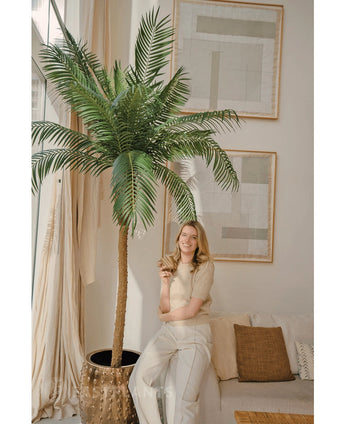 Een glimlachende vrouw staat naast de Kunstpalm Phoenix De Luxe 250 cm in een lichte, stijlvolle woonkamer met neutrale inrichting en grote abstracte kunst. Ze houdt een plantje vast en leunt nonchalant tegen een beige bank met kussens.