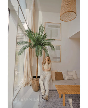 Een vrouw in lichtgekleurde kleding staat naast de Kunstpalm Phoenix De Luxe 250 cm in een lichte, moderne woonkamer met grote ramen, een beige bank, rieten lamp, houten salontafel en ingelijste muurkunst.