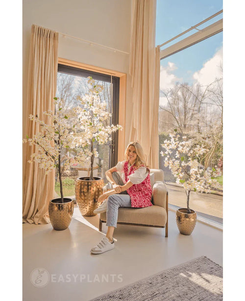 Una mujer se ríe en un sillón beige cerca de grandes ventanales con cortinas beige, rodeada por tres buganvillas artificiales de 150 cm blancas en un espacio luminoso, moderno y lleno de luz natural.