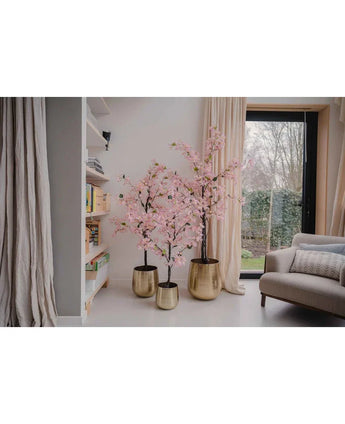 Een Kunstplant Bougainvillea 180 cm roze staat in een gouden plantenbak bij een raam in een moderne woonkamer, naast een boekenplank en beige gordijn, met rechts een grijze fauteuil zichtbaar.
