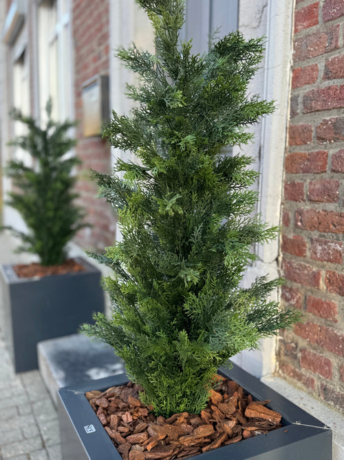 Un primer plano de la Planta Artificial Conífera UV 120 cm, una conífera artificial verde en una maceta rectangular negra con mantillo de corteza marrón, expuesta en una acera cerca de un edificio de ladrillo; otra jardinera de este tipo está al fondo.