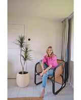 Una mujer con un top rosa brillante y vaqueros azules está sentada descalza en una moderna silla negra y de mimbre junto a la planta artificial Dracaena Fragnans de 150 cm en una habitación luminosa y minimalista con paredes y suelo blancos.