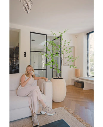 Una mujer con ropa de estar por casa clara está sentada en un sofá beige y sonríe con una taza de café en un moderno salón con suelos de madera, ventanales y el Enkianthus artificial de 180 cm como gran planta en maceta.