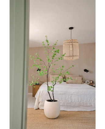 Un dormitorio minimalista con la planta Artificial Enkianthus de 180 cm a modo de gran maceta, una cama con ropa de cama blanca, una lámpara colgante tejida, paredes beige y una alfombra de fibras naturales para un ambiente sencillo y aireado.