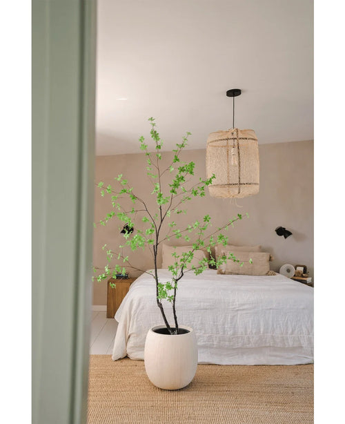 Un dormitorio minimalista con la planta Artificial Enkianthus de 180 cm a modo de gran maceta, una cama con ropa de cama blanca, una lámpara colgante tejida, paredes beige y una alfombra de fibras naturales para un ambiente sencillo y aireado.