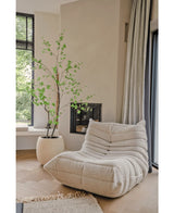 Un acogedor sillón con textura de color crema se encuentra cerca de una gran ventana y una chimenea. Cerca de allí, la planta artificial Enkianthus de 180 cm añade un toque natural de verde a esta habitación luminosa, aireada y moderna.