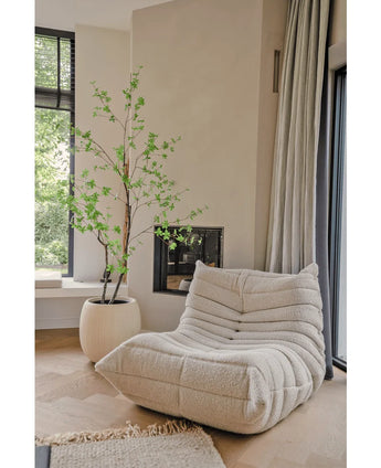 Un acogedor sillón con textura de color crema se encuentra cerca de una gran ventana y una chimenea. Cerca de allí, la planta artificial Enkianthus de 180 cm añade un toque natural de verde a esta habitación luminosa, aireada y moderna.