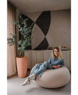 Een vrouw met krullend haar, gekleed in spijkerstof en witte sneakers, glimlacht terwijl ze op een ronde stoel zit. Achter haar staat de Kunstplant Eucalyptus 180 cm, een dressoir en abstracte muurkunst.