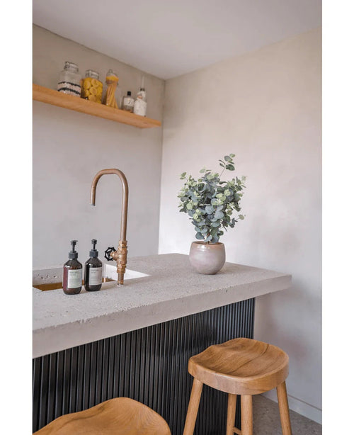 Una moderna encimera tiene un grifo de latón, dosificadores de jabón, la maceta de Eucalipto Artificial de 61 cm y dos taburetes de madera. Encima de la encimera, los estantes en una pared clara contienen frascos y botellas.