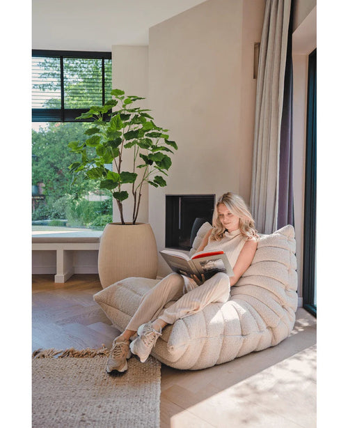 Una mujer lee en un salón moderno y luminoso, sentada en un acogedor sillón junto a la ventana, cerca de una planta artificial Ficus de 150 cm y suaves cortinas, rodeada de luz natural.