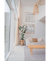 Een lichte, moderne woonkamer met grote ramen, een witte bank, neutrale kussens, een geweven salontafel, twee abstracte muurkunstwerken, een Kunstplant Heliconia 180 cm in een pot met structuur en geweven hanglampen die aan het plafond hangen.