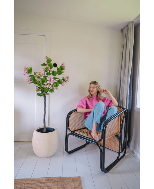 Una mujer con un jersey rosa y vaqueros azules está sentada descalza en una silla de ratán junto a la Planta Artificial Judas 150 cm, un árbol artificial con flores rosas, en una habitación moderna y luminosa con paredes y suelos blancos.
