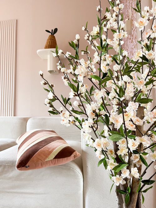 Una acogedora sala de estar con un sofá blanco, un cojín a rayas rosas y marrones y en primer plano la rama floreciente de cerezo artificial color crema de 120 cm. Una planta en maceta descansa sobre un estante de pared contra una pared de color rosa pálido.