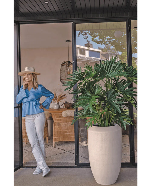 Una mujer con blusa azul, pantalón blanco y sombrero se ríe ante una puerta de cristal junto a la planta Filodendro artificial de 125 cm en una maceta beige. Las sillas y la decoración de mimbre son visibles en toda la habitación.