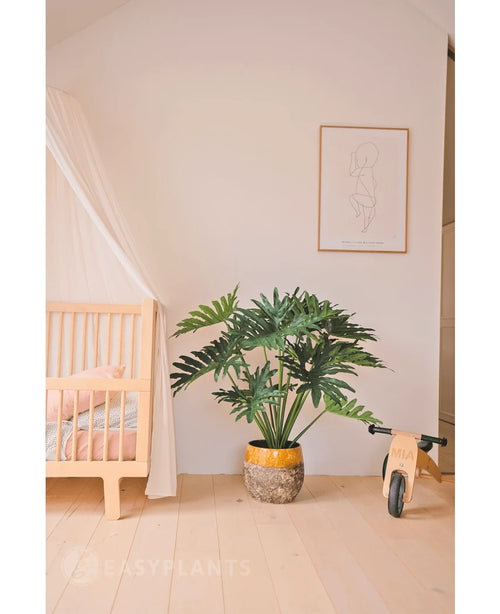 Una habitación infantil acogedora y minimalista con una cuna y un dosel de madera a la izquierda, una planta artificial de filodendro UV de 90 cm en el centro como obra maestra, a la derecha una bicicleta de juguete de madera y encima una sencilla impresión artística lineal.