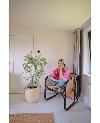 Een vrouw in een roze shirt en spijkerbroek zit blootsvoets op een geweven stoel, glimlachend en ontspannend in een lichte, minimalistische kamer met witte muren en vloeren. Naast haar staat de Kunstplant Ruscus 120 cm.
