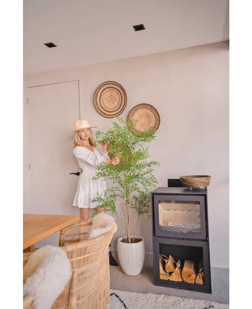 Una mujer con un vestido blanco y un sombrero está junto a la planta Ruscus artificial de 150 cm, que aporta un toque natural a una habitación moderna con muebles de mimbre, cestas de pared y una estufa de leña negra, perfecta para decorar interiores.