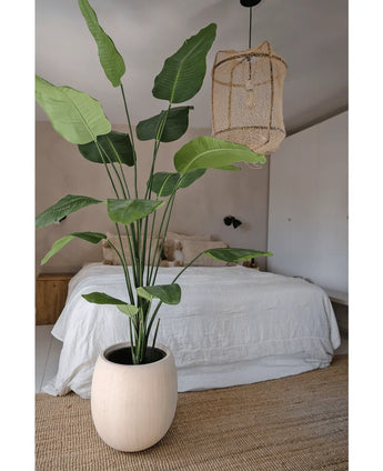 De Kunstplant Strelitzia 210 cm real touch met hoge groene bladeren staat op een geweven vloerkleed in een neutrale slaapkamer, met een knus bed met wit beddengoed en een rieten hanglamp erboven.