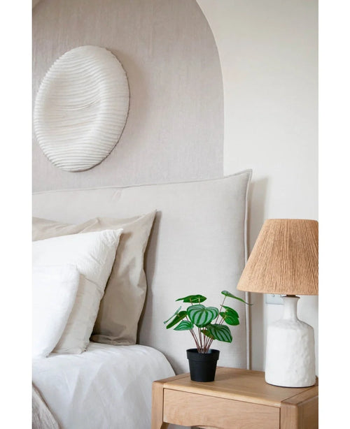 Un acogedor dormitorio dispone de una cama con ropa de cama neutra, una mesita de noche de madera clara con la planta de Sandía Artificial de 29 cm y una lámpara blanca con pantalla de mimbre. Una escultura de pared con textura blanca cuelga encima de la cama.