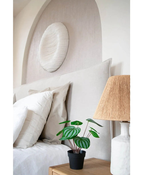 Una cama cuidadosamente hecha con almohadas y ropa de cama blancas se encuentra junto a una mesita de noche de madera con la planta de Sandía Artificial de 29 cm y una lámpara con pantalla tejida; encima de la cabecera cuelga una escultura de pared con textura blanca.