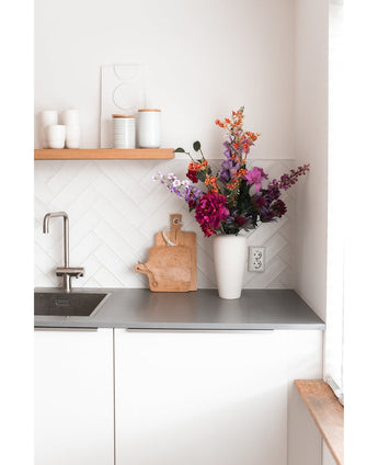 Una cocina blanca moderna con una encimera gris, un fregadero, una tabla de cortar de madera y un jarrón blanco lleno de Hortensias Artificiales de 55 cm de color rosa oscuro. Tazas y ollas blancas se encuentran sobre un estante de madera encima del mostrador.