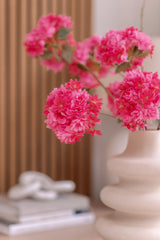 Un jarrón de cerámica blanca sostiene una rama artificial de cerezo en flor de 85 cm de color rosa. El fondo muestra paneles verticales de madera, una pila de libros y un pequeño objeto decorativo encima.
