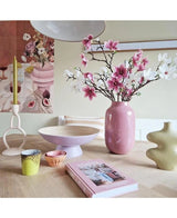 Una habitación moderna tiene una mesa con rama artificial de magnolia de flores pequeñas de 89 cm de color rosa en un jarrón rosa, candelabros, cuencos, tazas de colores pastel, un jarrón escultórico verde, un libro rosa y obras de arte florales en la pared.