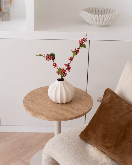 La rama de bayas artificiales rosa de 64 cm, un jarrón blanco lleno de ramas de bayas artificiales de color rosa y hojas verdes, se encuentra sobre una pequeña mesa redonda junto a una silla beige con un cojín marrón en una habitación acogedora y minimalista.