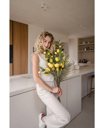 Una mujer de pelo rubio rizado, vestida con un traje de color crema, se apoya en un mostrador y sostiene el ramo 'Artificial Branch Lemon 104 cm yellow', decorado con exuberante vegetación. La moderna cocina está decorada en colores neutros y tiene detalles en madera.