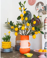 Una habitación luminosa con un jarrón alto lleno de limones y hojas verdes, en el que ocupa un lugar destacado el llamativo Kunsttak Lemon 104 cm amarillo, rodeado de cuencos de cerámica de colores, un cuenco con limones y platos decorativos en la pared, creando un aspecto alegre y ecléctico.