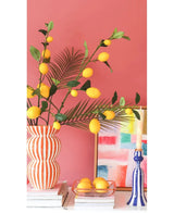 Contra una pared rosa coral hay un jarrón a rayas lleno de ramas de Kunsttak Lemon de 104 cm en amarillo, un cuadro colorido, una pila de libros, un candelabro azul con una vela y una bandeja de limones.